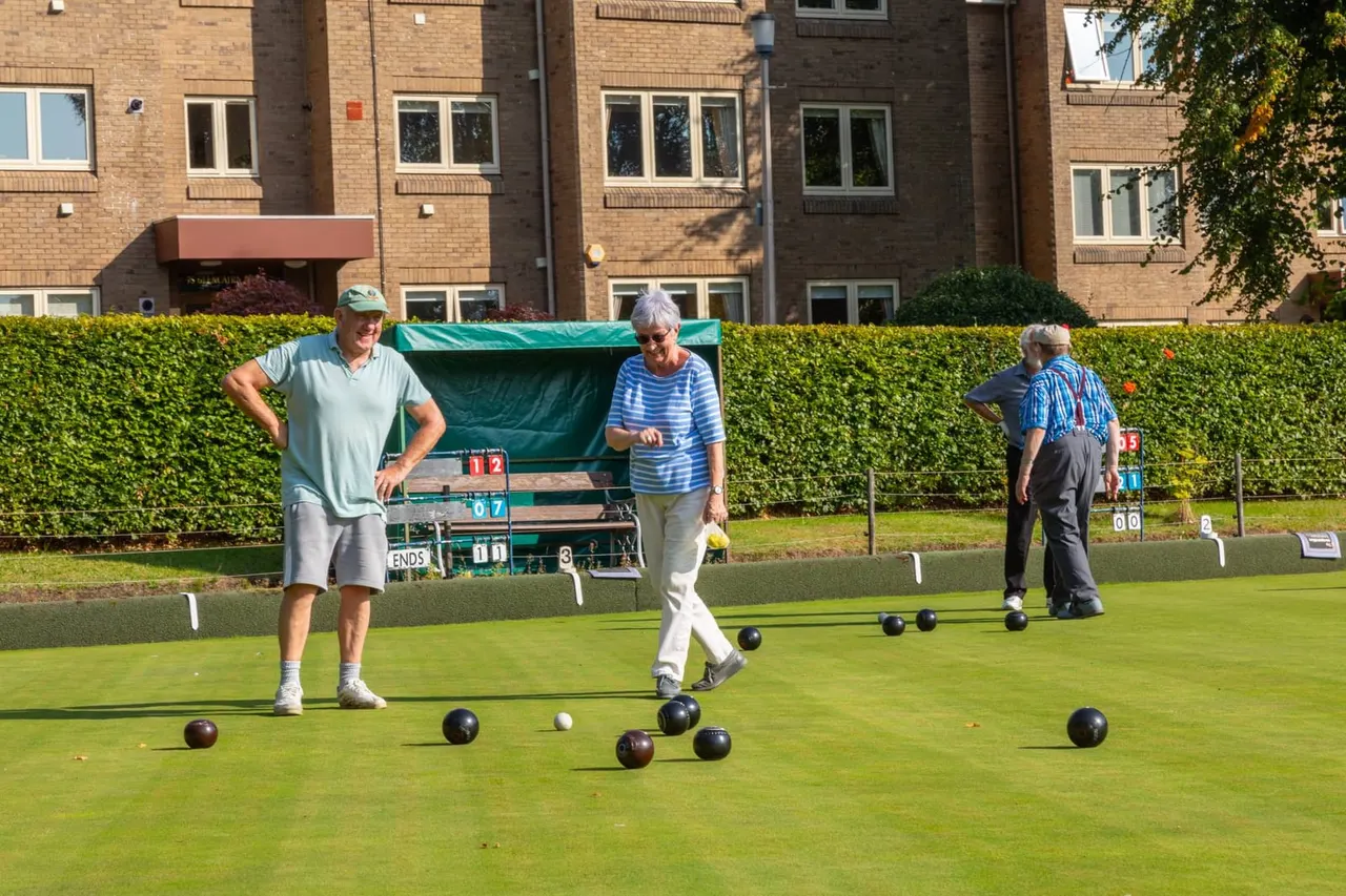Older adults playing bowls on a manicured green, with scoring equipment and apartment buildings visible beyond the hedged boundary.