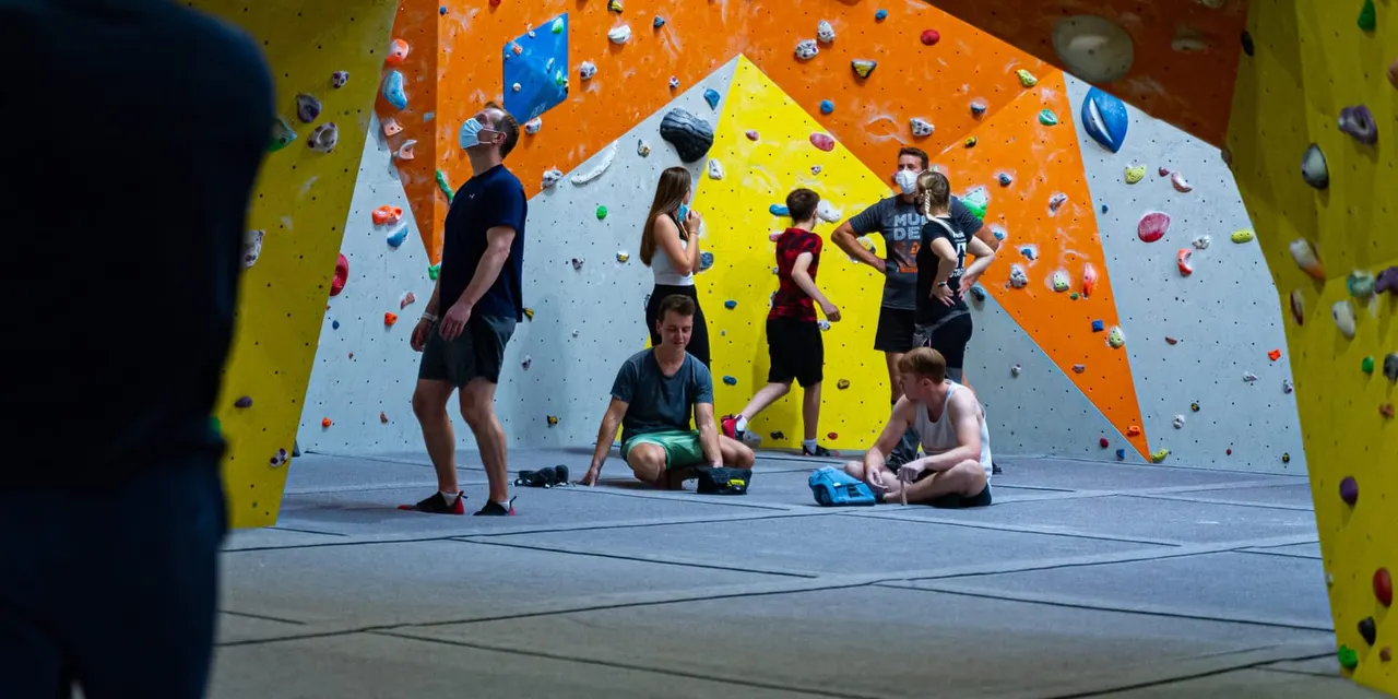A group of children and adults on a padded floor in front of colourful indoor climbing walls with various hold points.