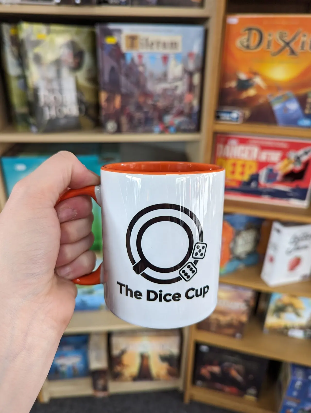Hand holding a white mug with orange rim branded "The Dice Cup" in front of board game boxes on shelves.