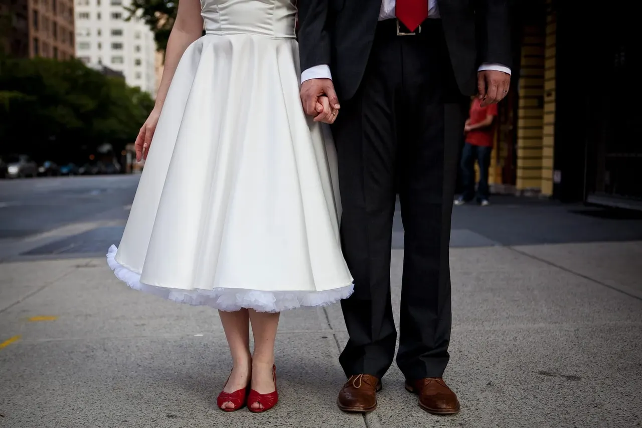 Bride in white dress and red shoes holds hands with groom in black suit on urban street.