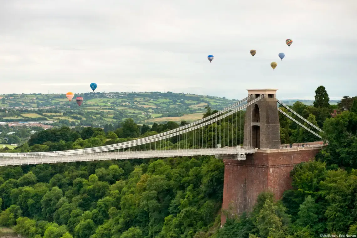 Multiple hot air balloons floating in the sky above the Clifton Suspension Bridge, with wooded gorge and rolling countryside below.