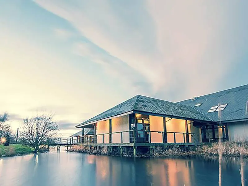 Modern waterside visitor centre with illuminated windows reflected in still water at dusk.