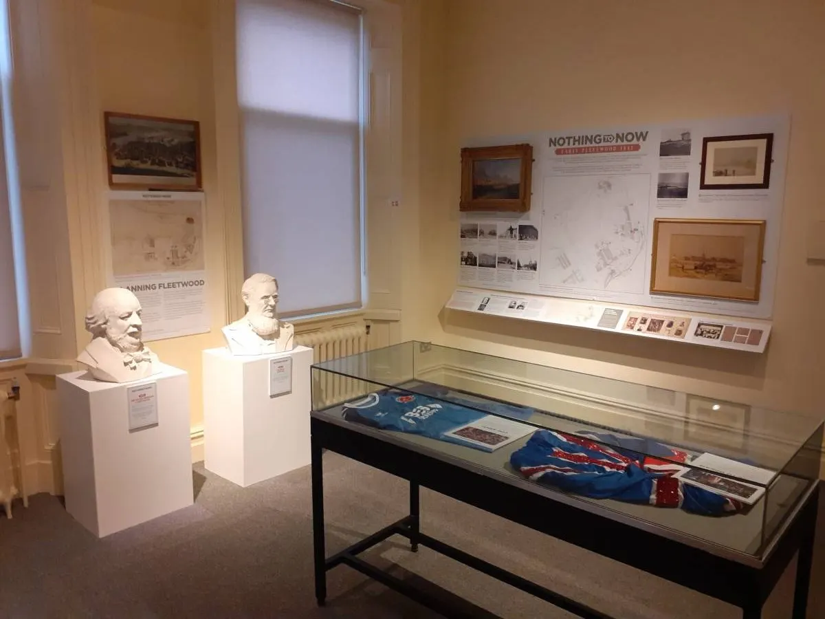 Museum display room with historical busts, framed artwork and maps on walls, and a glass display case containing artefacts and textiles.