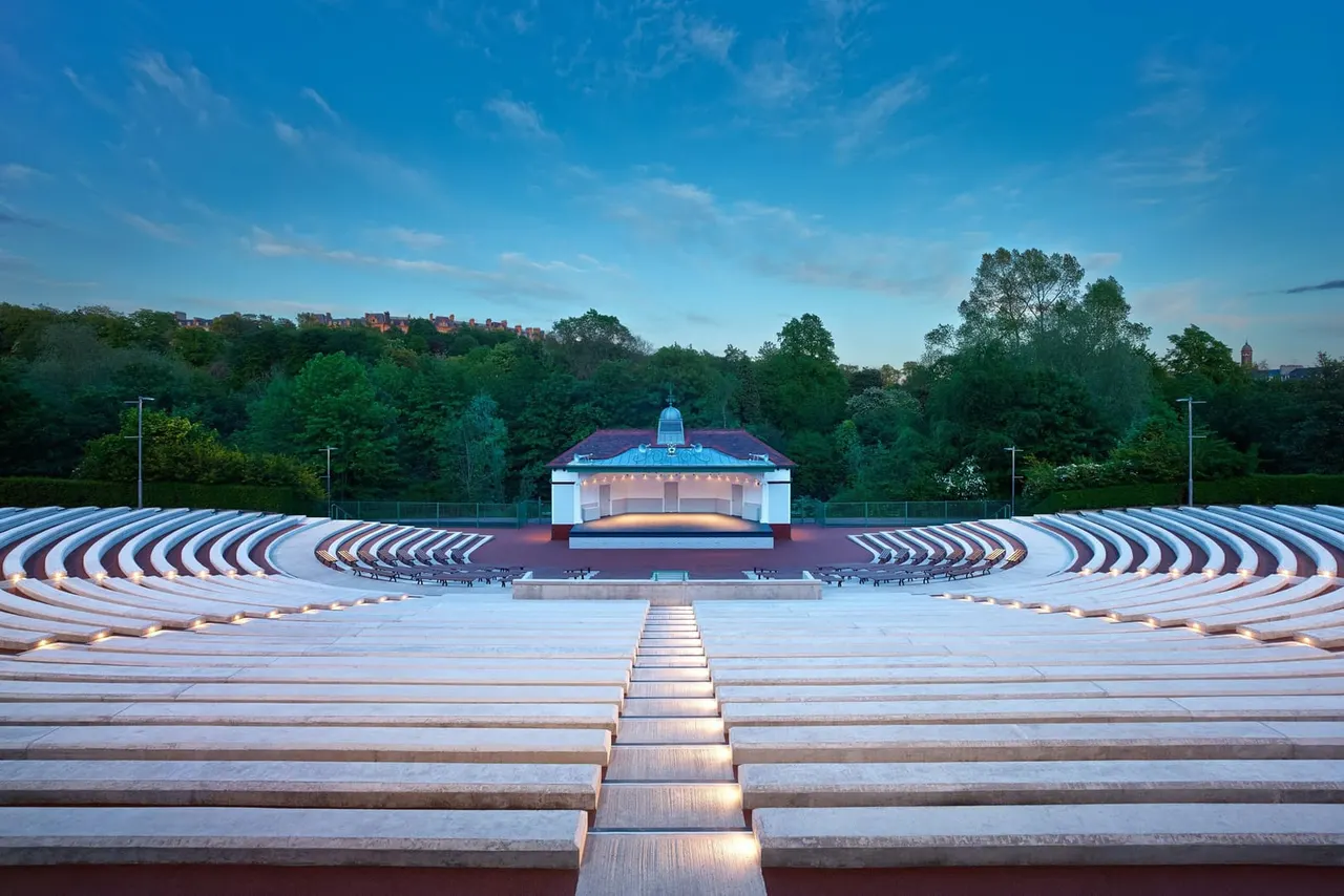 Empty amphitheatre with curved seating facing a lit bandstand, surrounded by trees under blue sky.