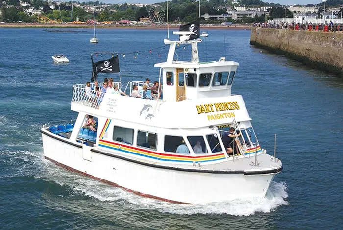 White passenger boat with pirate flag sailing in blue water with coastal town and pier visible behind.