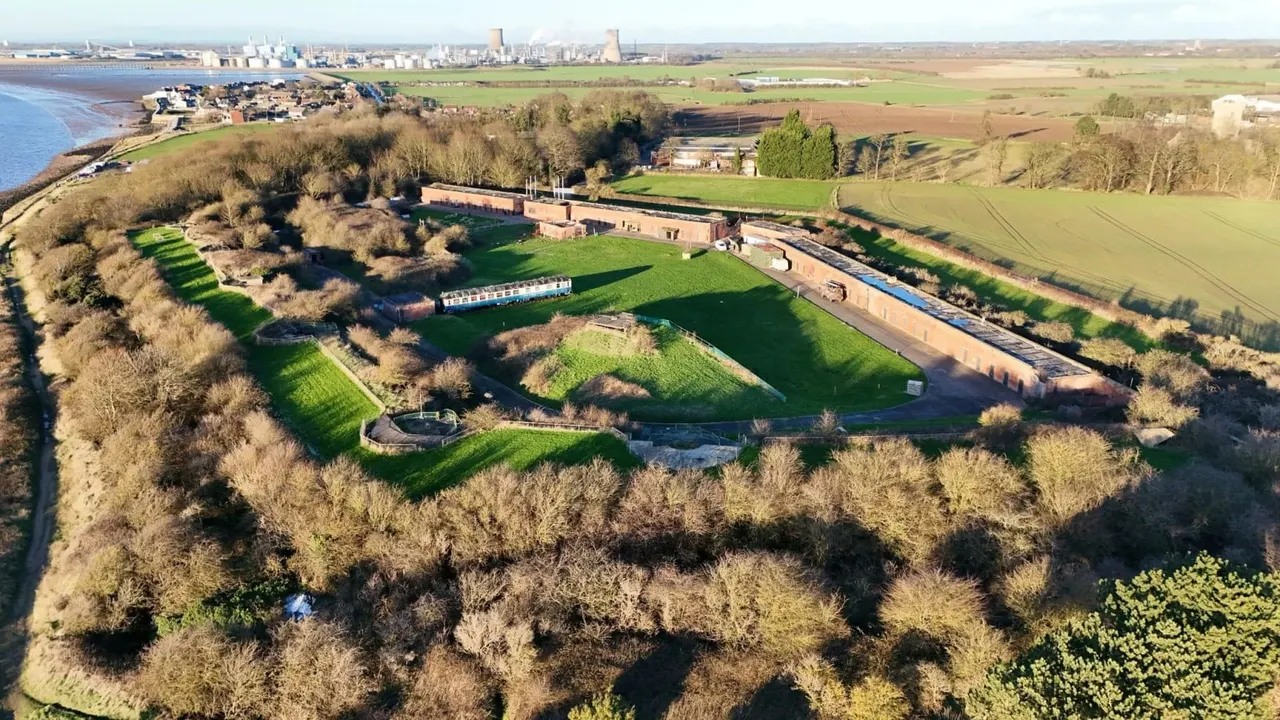 Aerial view of a waterside fort with green lawns, buildings and moored narrowboat surrounded by woodland, with industrial buildings and coastline beyond.