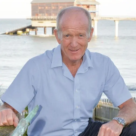 Man in light blue shirt smiling at camera by wooden pier with boathouse across the water.