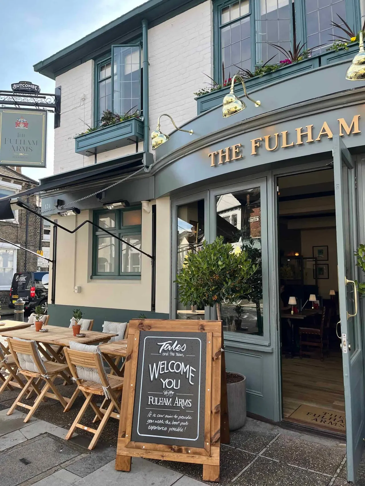 Exterior of a pub with outdoor seating, showing wooden tables, chairs, potted plants, brass hanging lamps and a welcome sign.