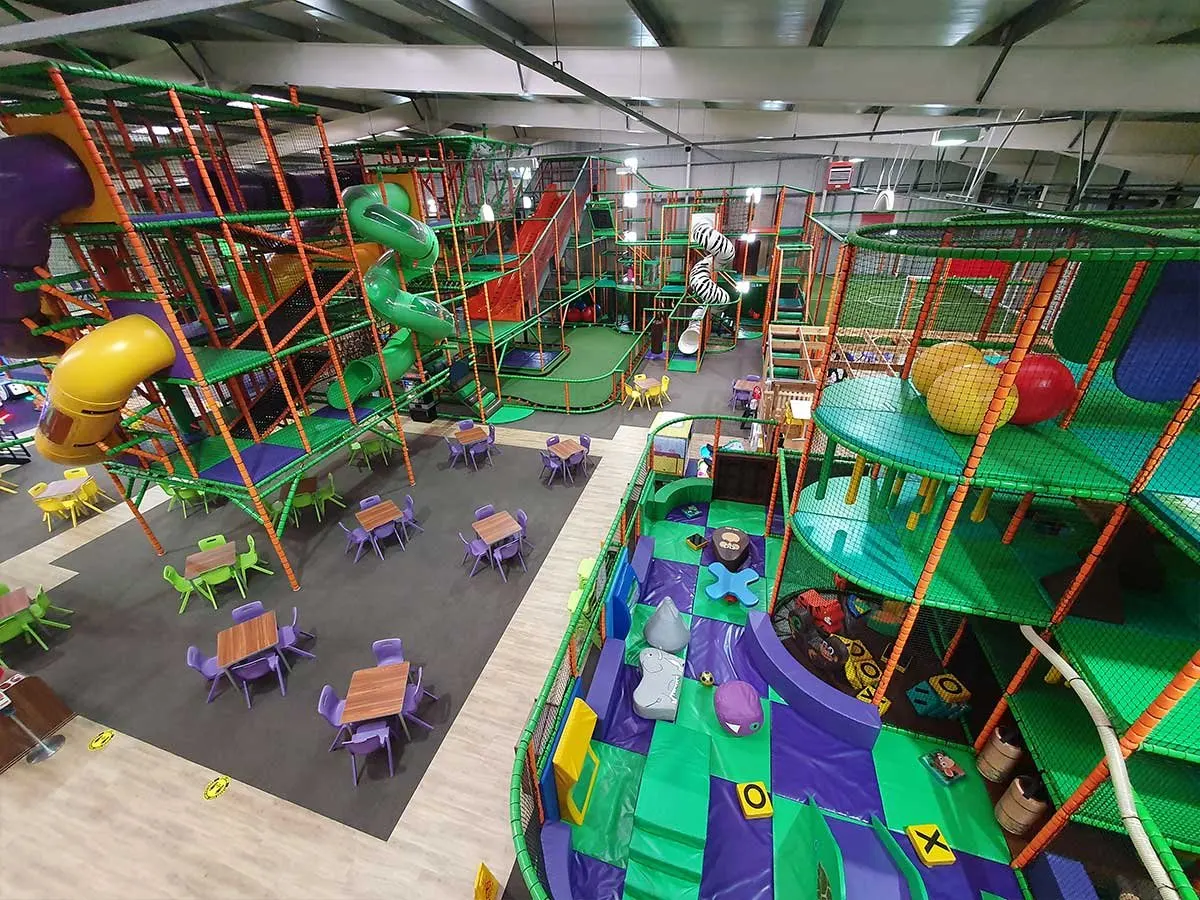 Indoor play centre with colourful climbing structures, slides, and soft play equipment, with seating areas and small tables below.