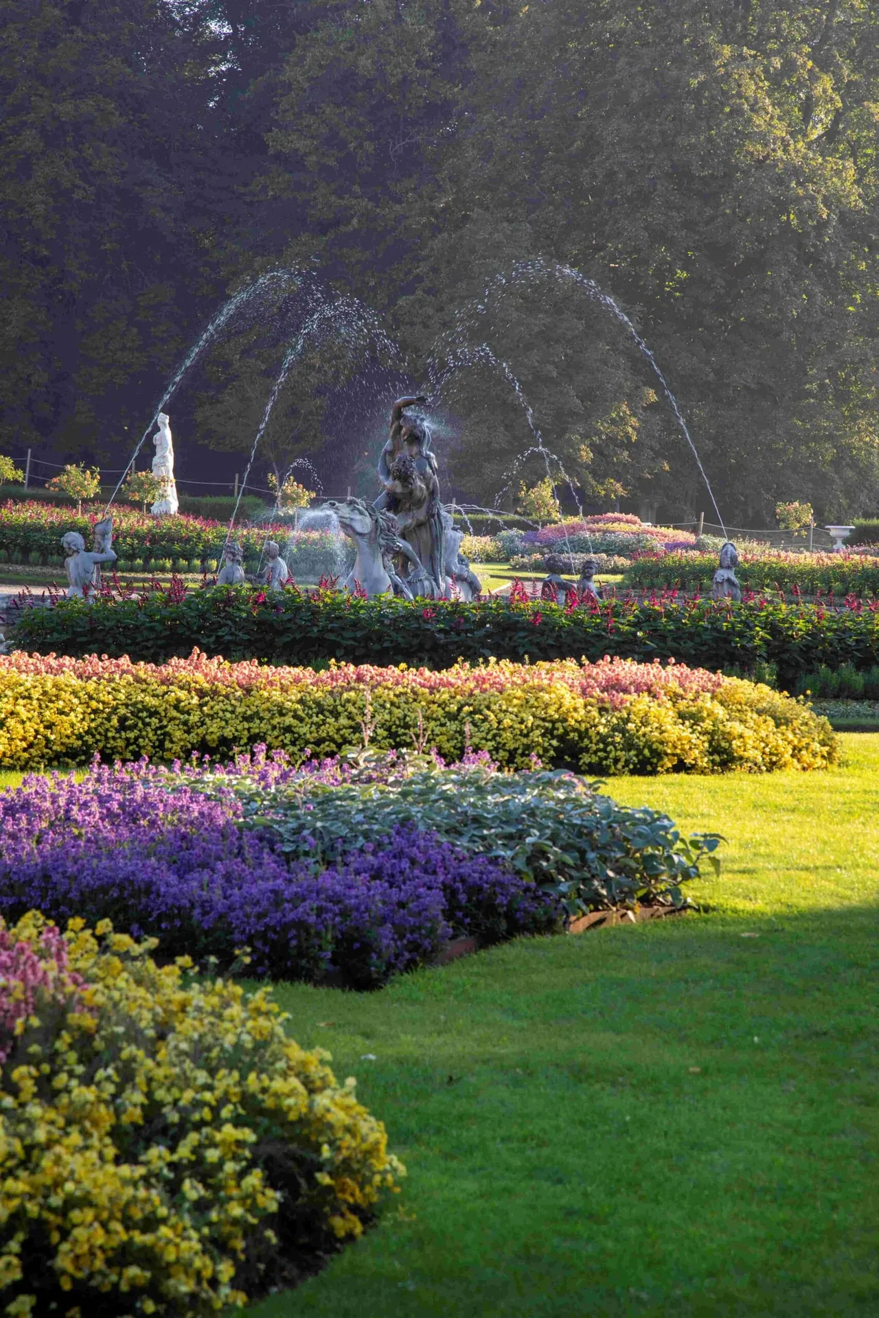 Formal gardens with colourful flower beds and hedges surrounding a fountain with arched water jets and stone statuary.