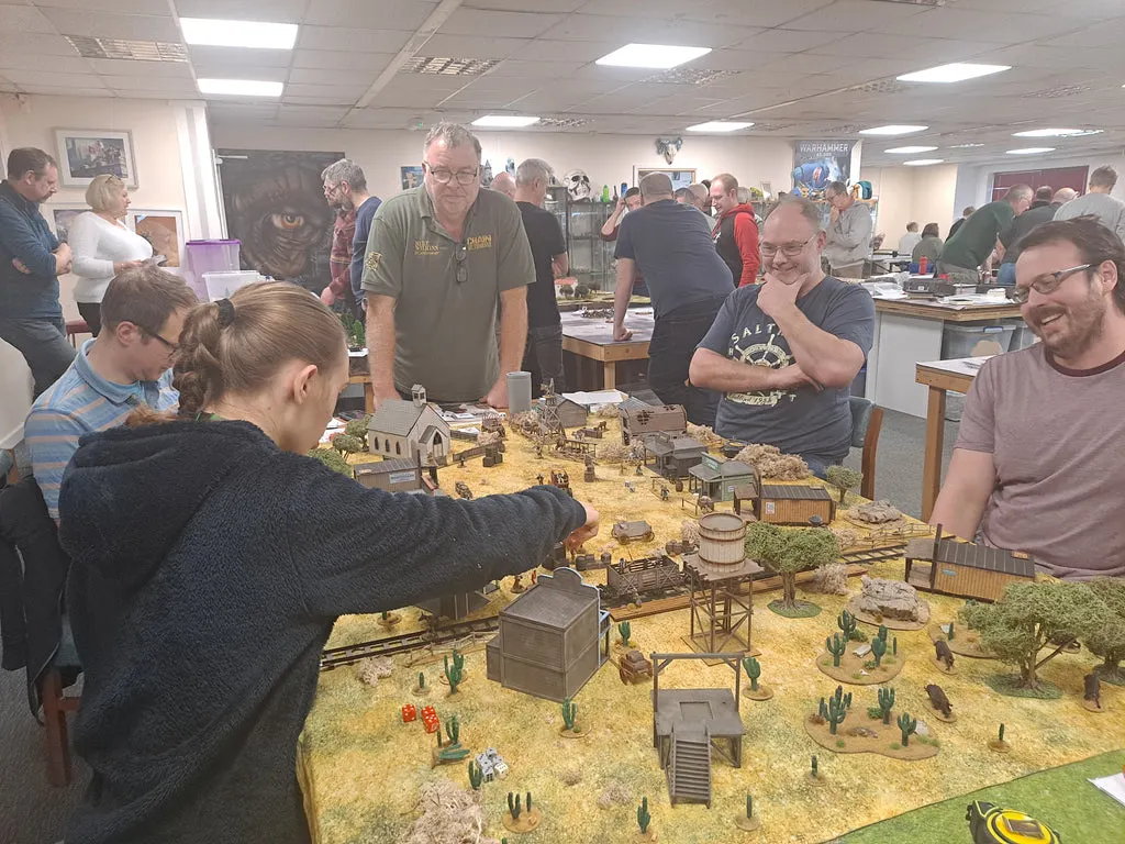 Young woman reaching towards a detailed miniature Western town diorama surrounded by spectators in an indoor gaming space.