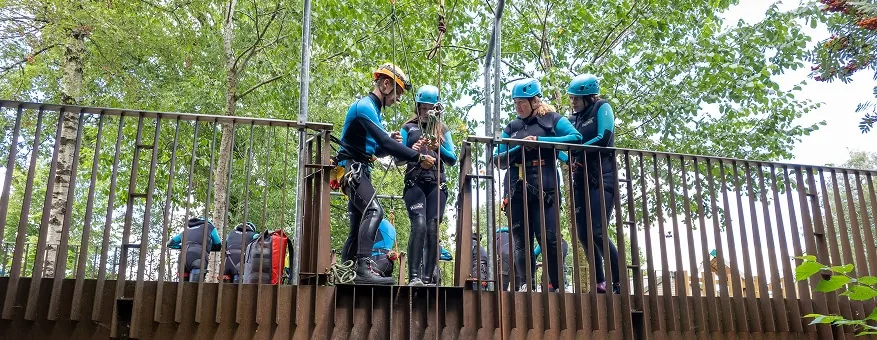 A group of people wearing blue hard hats and safety gear stand on a wooden platform amongst trees, preparing for an activity.