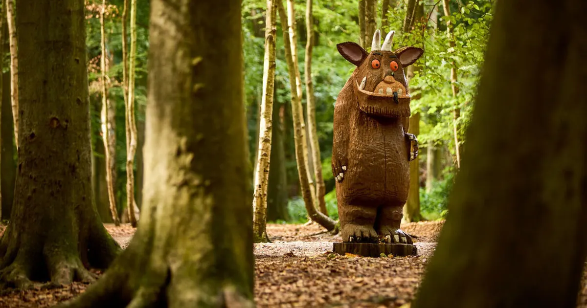 Carved wooden sculpture of a creature with horns and fangs standing on a forest path surrounded by moss-covered trees.