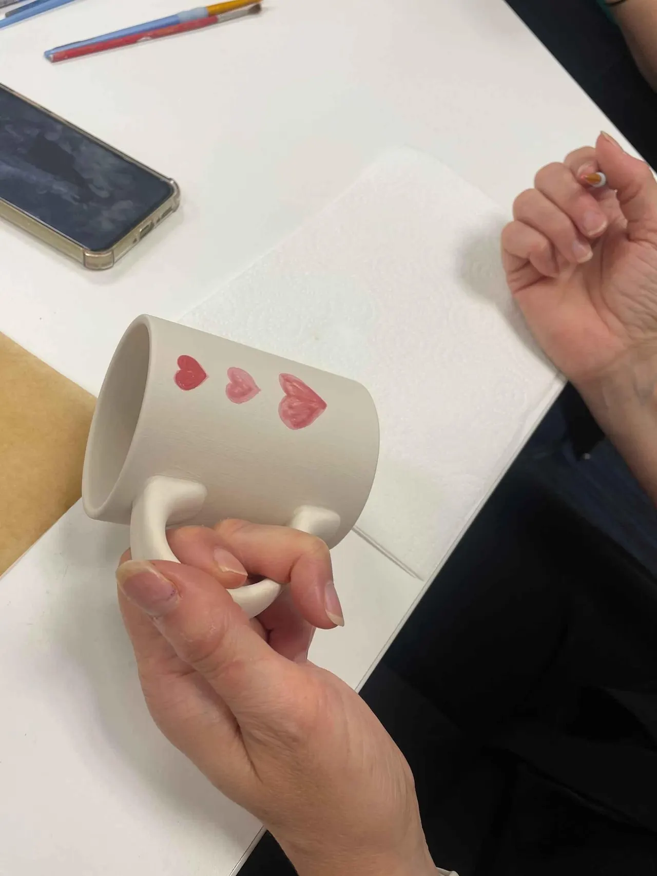 Child's hands holding a white ceramic mug decorated with pink hearts during a pottery painting activity.