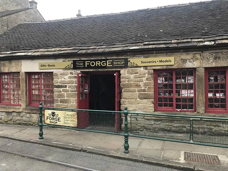 Stone building housing The Forge Shop with red-framed windows and doors, green railings and bollards, and tram tracks visible in foreground.