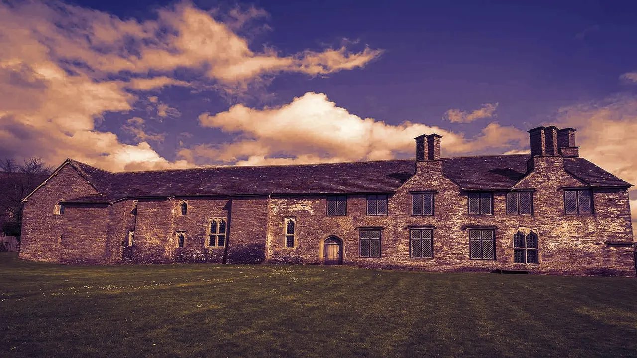 Historic stone manor house with multiple chimneys set against a dramatic purple and golden sky with white clouds.