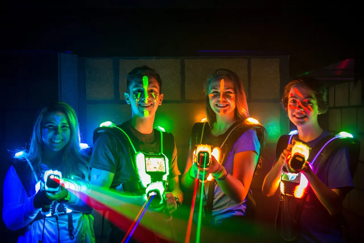 Five young people holding glowing laser tag weapons, illuminated by multicoloured neon lights in an indoor arena.
