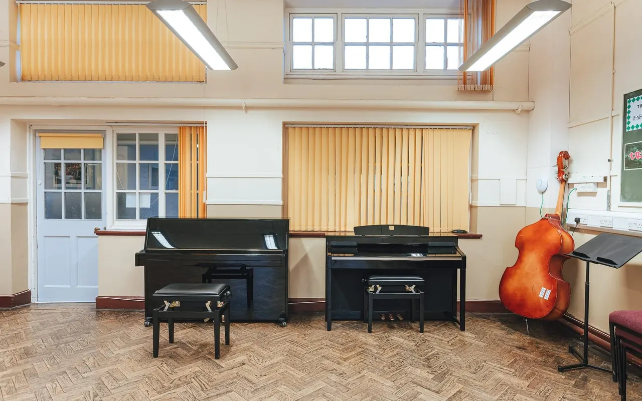 Music practice room with two black pianos, a cello, and music stands in a bright studio with wooden flooring and yellow vertical blinds.