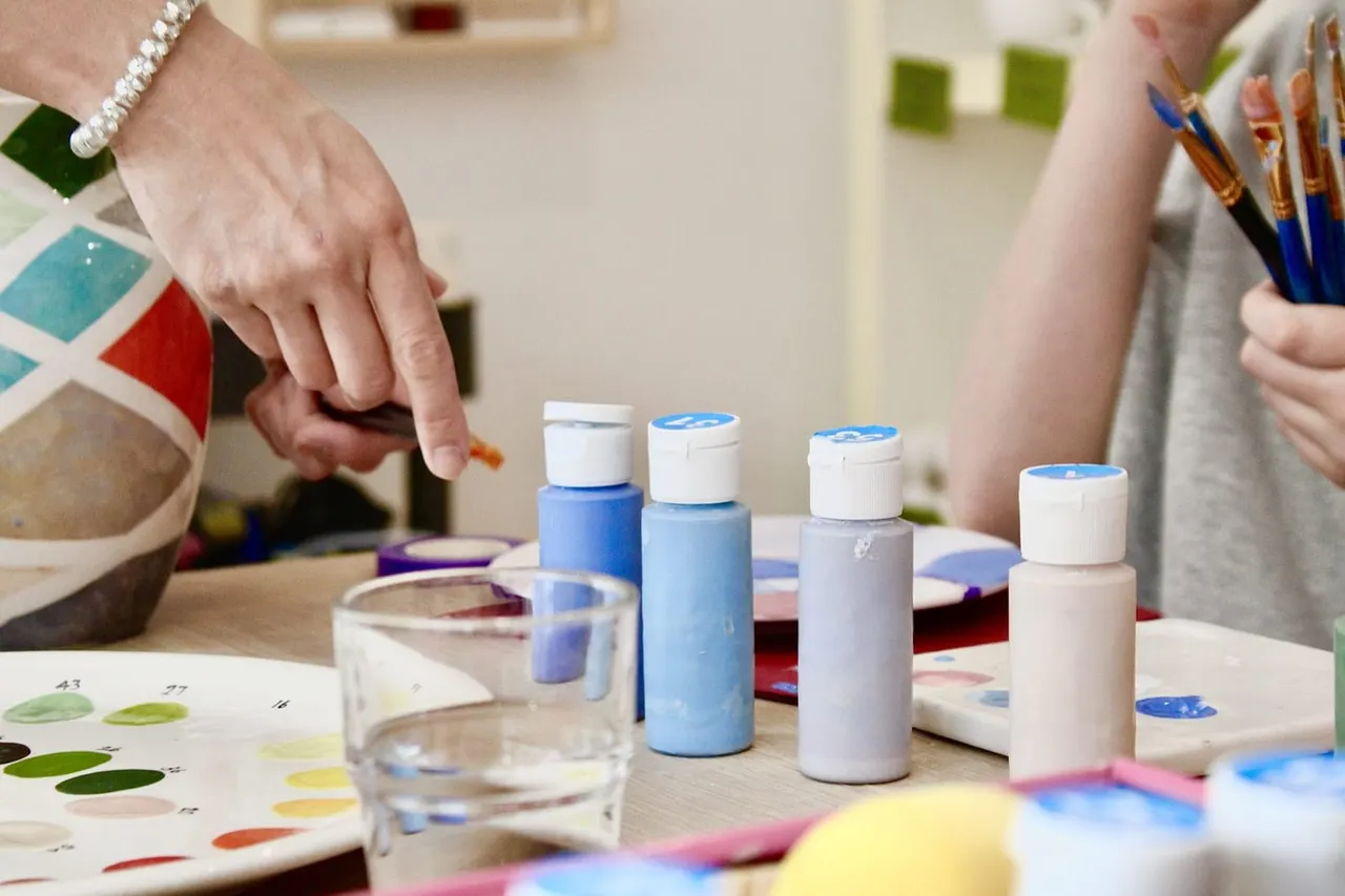 Person with pearl bracelet reaching toward blue and white paint bottles during a pottery painting activity.