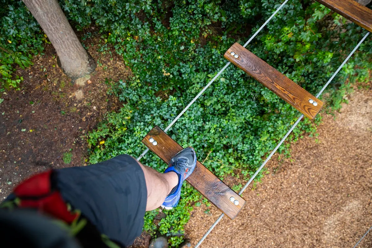 Person in blue trainers stepping on wooden rungs of a rope bridge suspended above green foliage canopy.