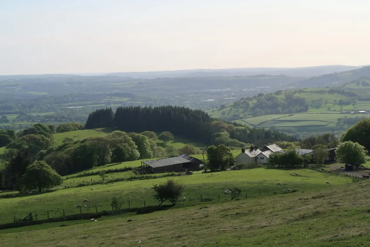 Rural farmstead nestled in rolling green countryside with farm buildings, houses, and expansive valley views beyond.