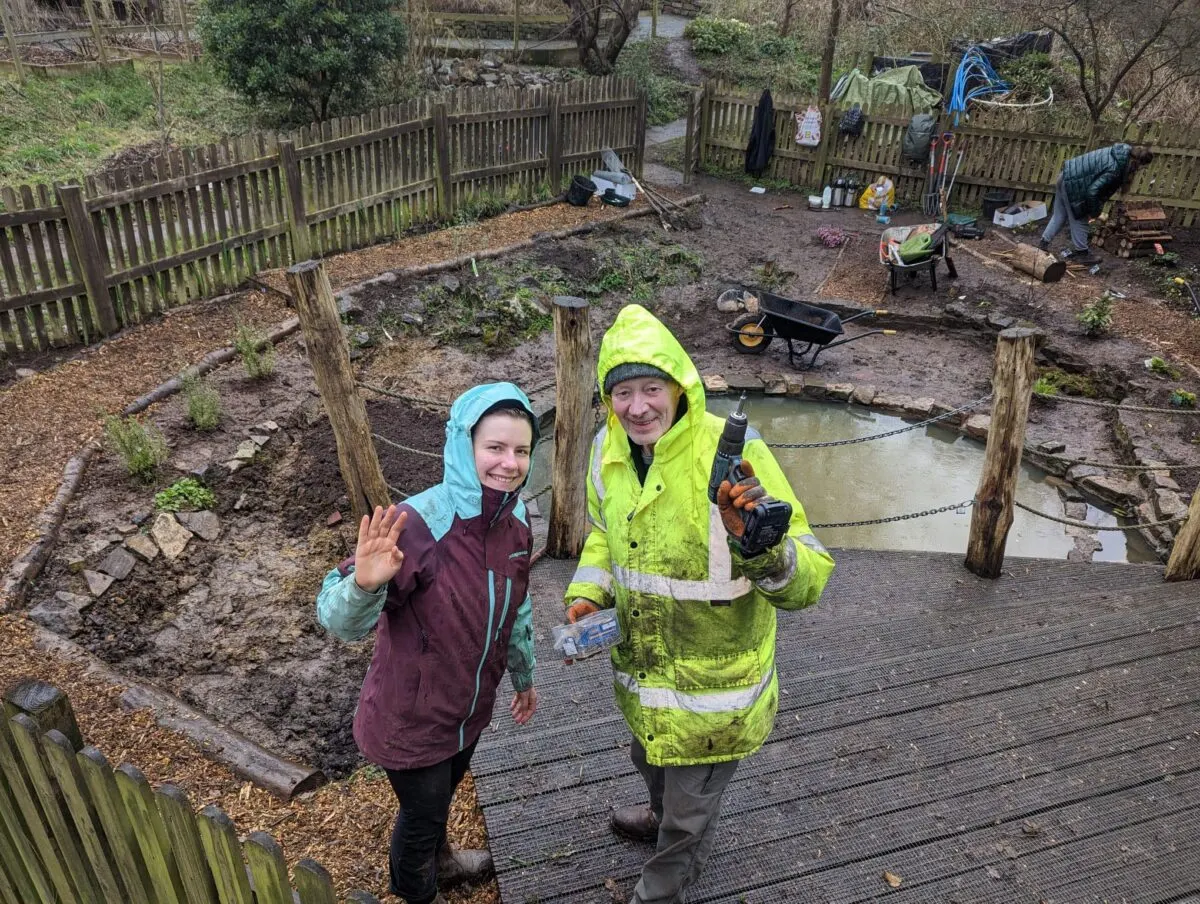 A child and adult in bright waterproof jackets stand on a wooden walkway beside a pond in a fenced community garden area.
