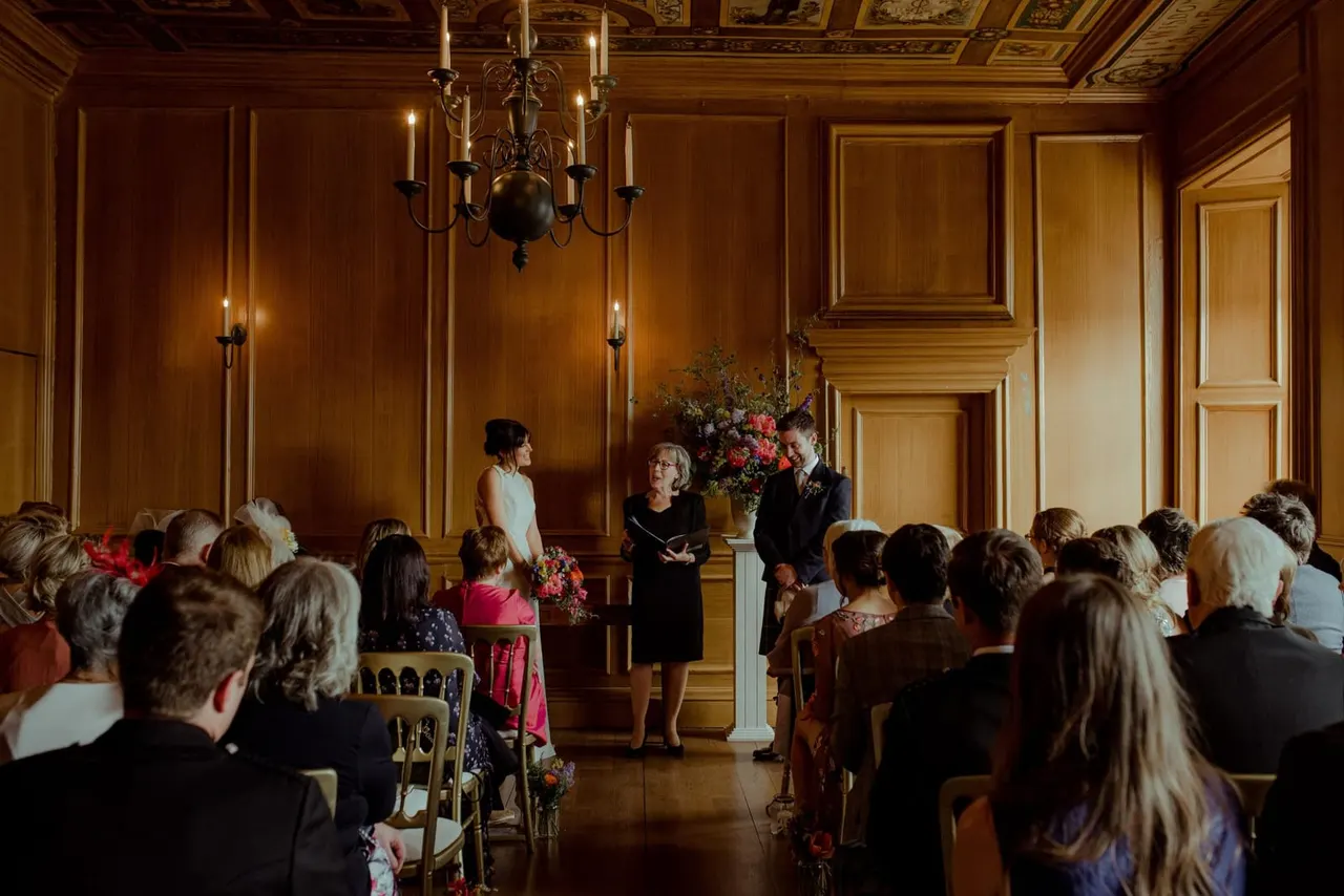 Ceremony taking place in an elegant wood-panelled room with guests seated facing a woman officiating between floral arrangements.