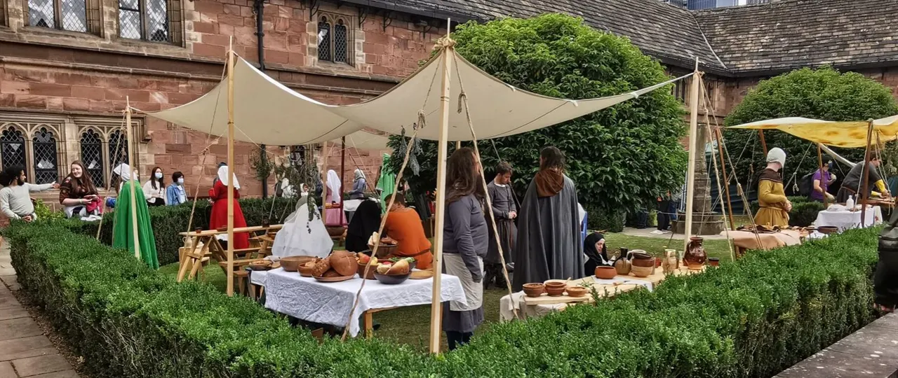 Medieval fair event in courtyard with visitors in period costume under canvas awnings beside historic stone building.