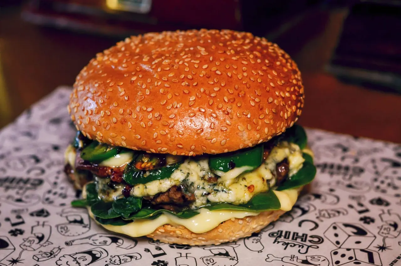 Gourmet burger with sesame seed bun, melted cheese, leafy greens and seasoned patty on patterned paper wrapper.