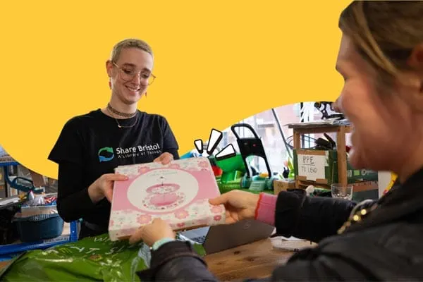 Two people exchange a pink boxed item at a wooden table in front of a bright yellow background, surrounded by shared goods and supplies.