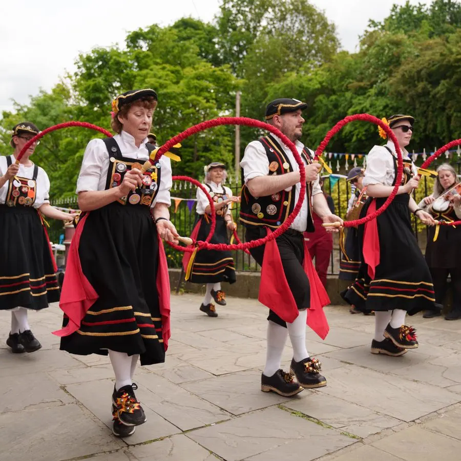 Performers in traditional folk costumes dancing with red hoop props outdoors in a garden setting.