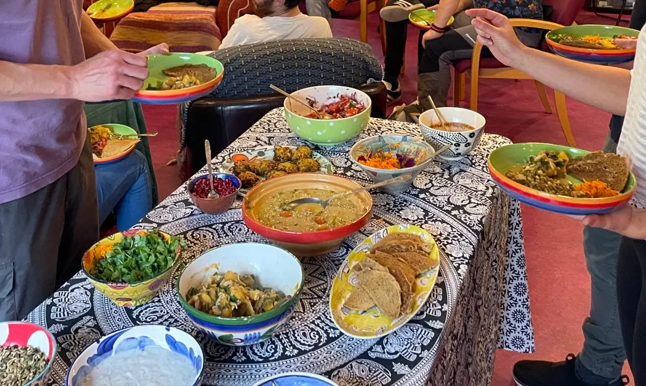 A group of people seated around a table sharing a spread of Middle Eastern or South Asian dishes including hummus, flatbread, salads, and curries.
