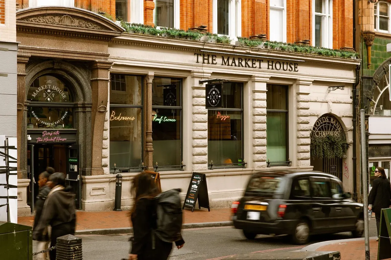 Historic stone building with Market House signage, street-level shopfronts advertising bedrooms and dining, pedestrians walking past, black taxi cab on street.