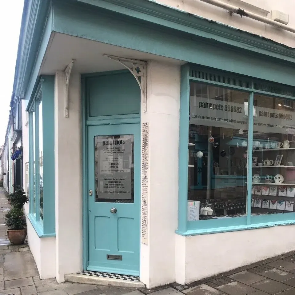 Exterior of a small turquoise and cream shopfront with a corner entrance door and display windows showing craft supplies and pottery items.