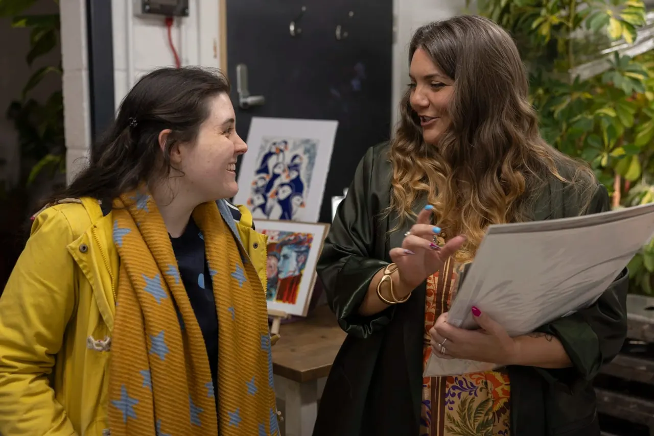 Two women converse in an art gallery, one holding a tablet, with framed artworks visible on the wall behind them.
