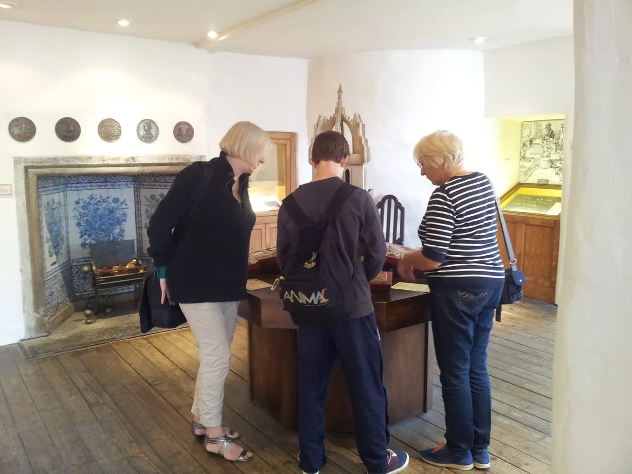 Three visitors exploring a museum room displaying historical artefacts, coins and a decorative fireplace.