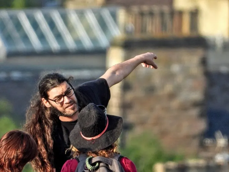 A guide with long hair and glasses points whilst holding a large bird, with a child beside them and a stone building in the background.