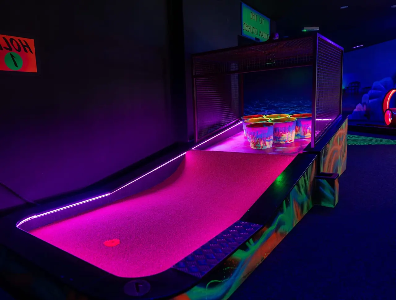 A neon-lit indoor mini golf hole with vibrant pink and purple lighting and colourful cups and obstacles.