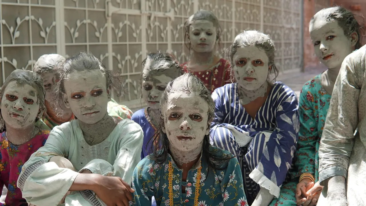 Group of children with faces covered in white powder or clay, wearing colourful patterned clothing, standing together outdoors against a decorative metal gate.