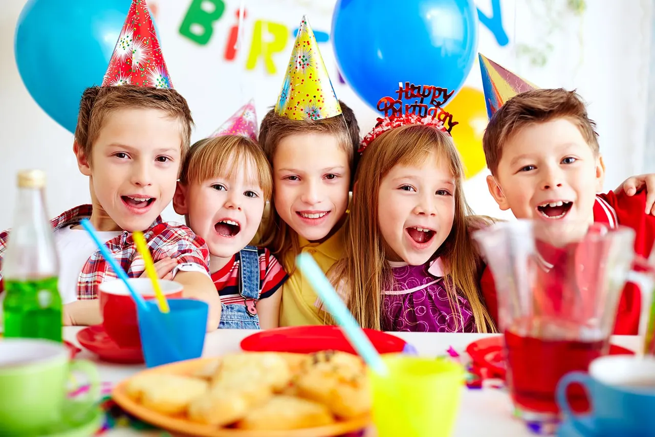 Five children wearing colourful party hats laugh at a birthday party table with balloons, drinks and food.