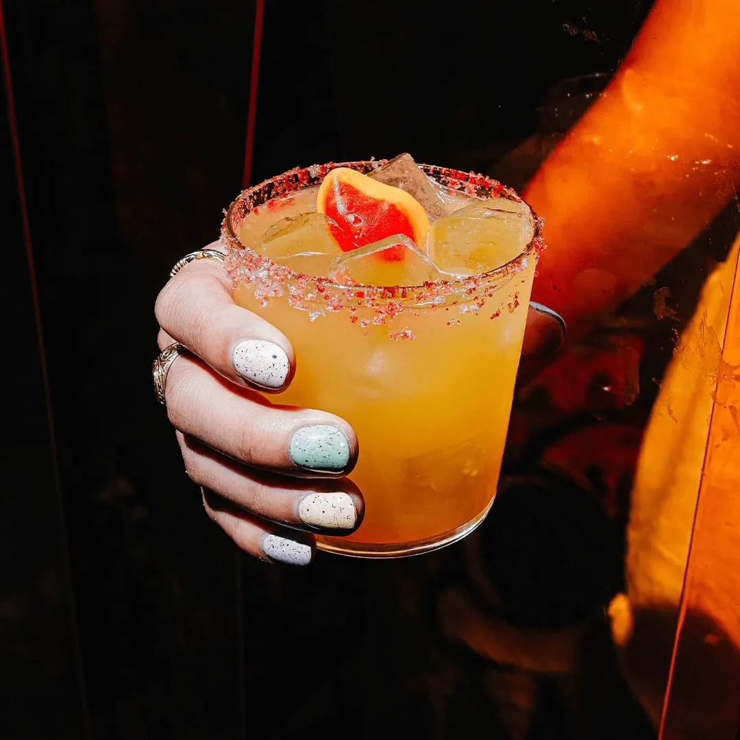 Hand holding a colourful cocktail with a spiced sugar rim and grapefruit slice garnish against a dark background.