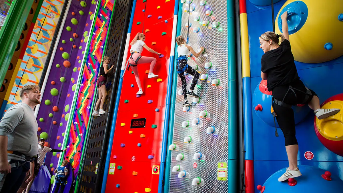 Multiple people of various ages climbing coloured indoor climbing walls with handholds and safety equipment.