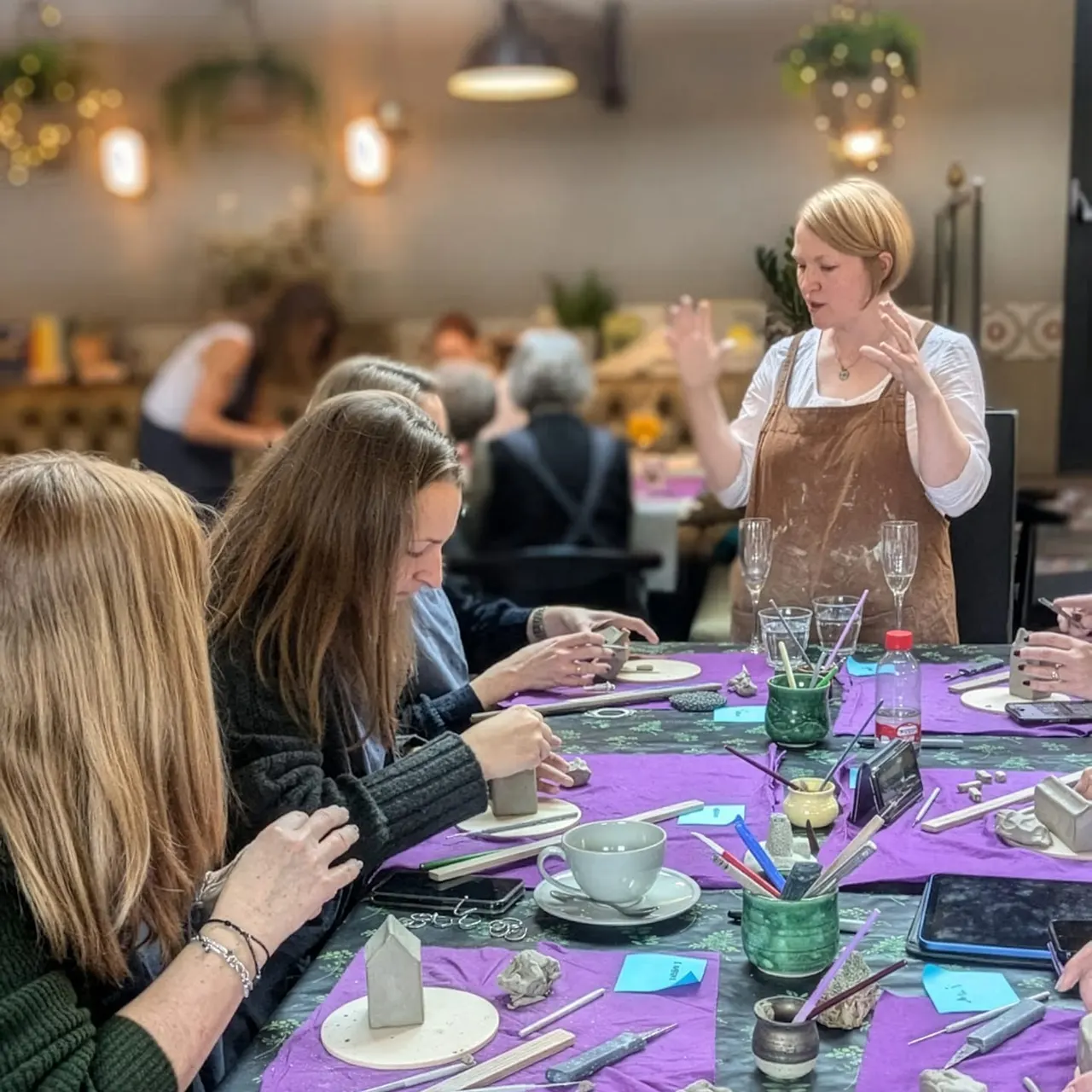 An instructor demonstrates a pottery technique to participants seated at a purple work table during a creative workshop.