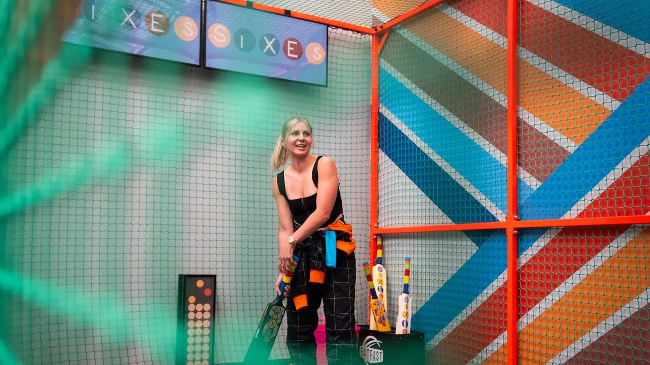 Woman holding a cricket bat stands in a colourful indoor cricket booth with netting and a digital scoreboard display.