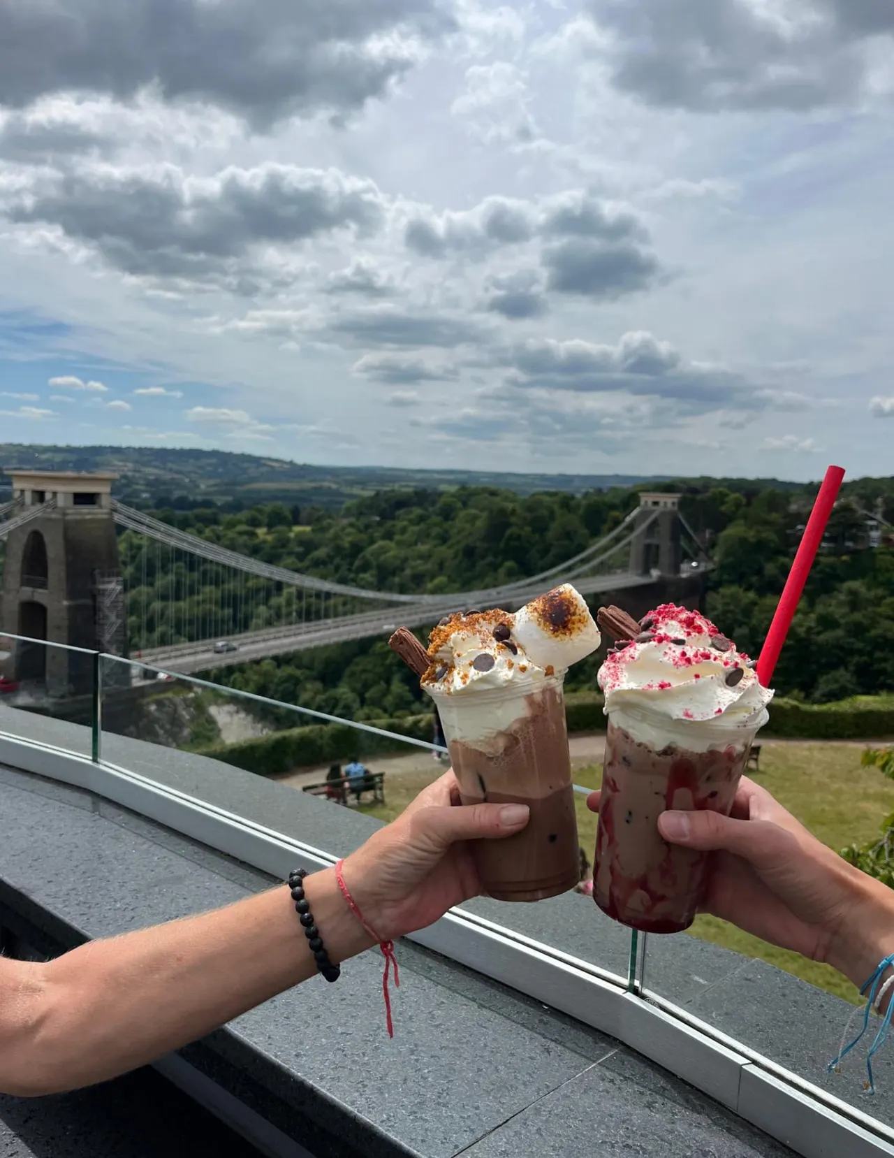 Two hands holding decorated milkshakes with whipped cream and toppings, with Clifton Suspension Bridge and wooded valley visible beyond.