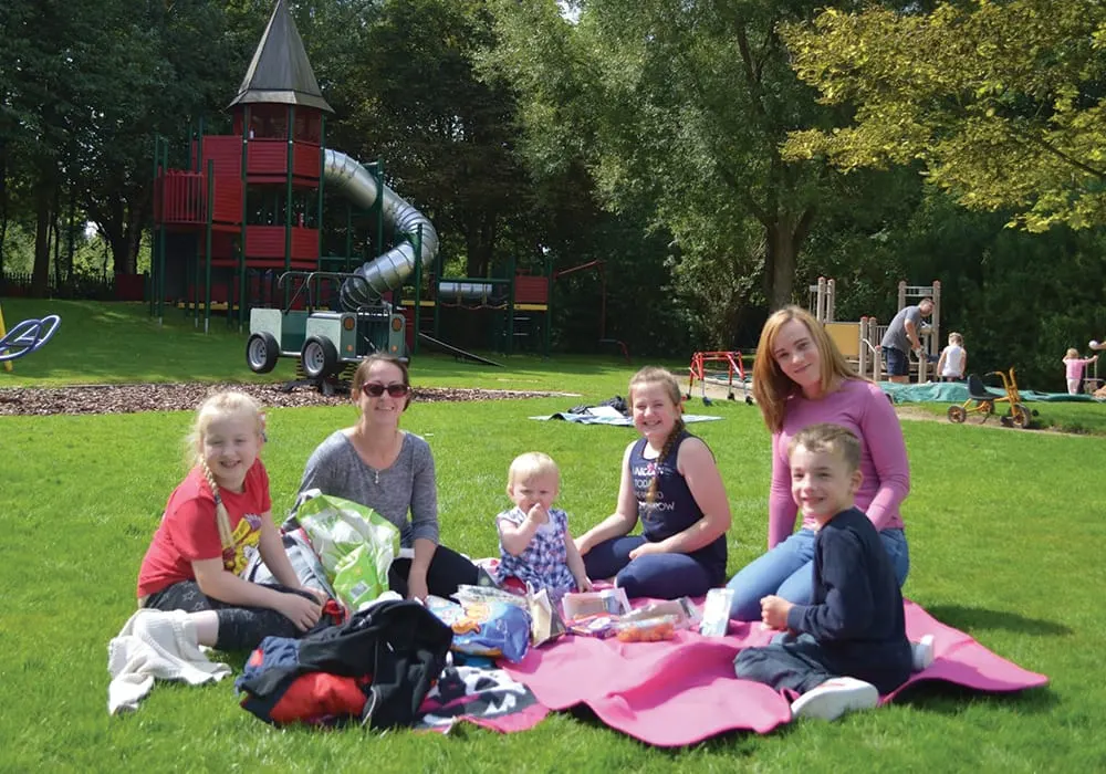 Adults and children having a picnic on grass with a red castle-themed playground structure and slide behind them.