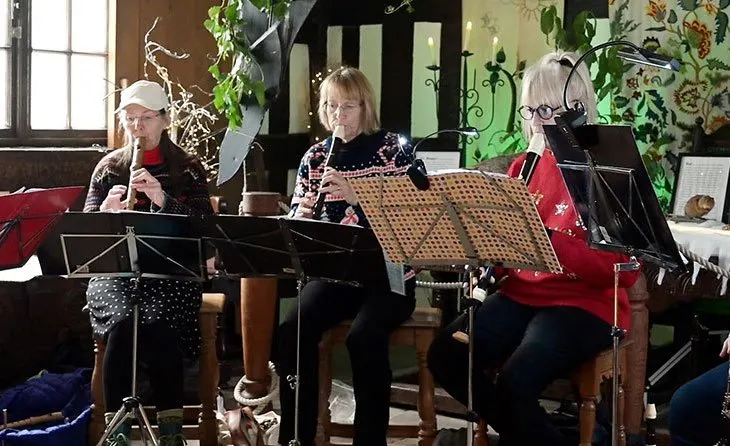Three older women perform traditional music together indoors, playing wind instruments and reading from sheet music on stands.