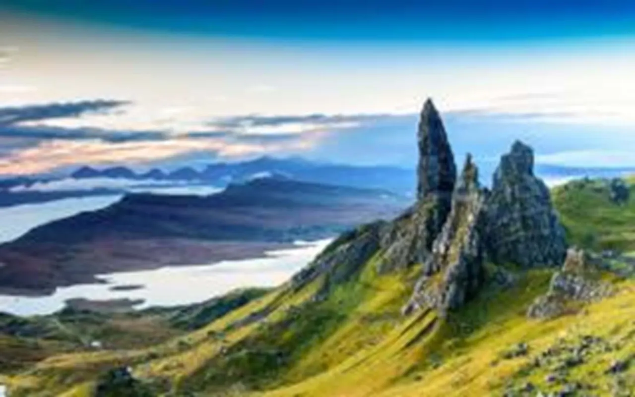 Elevated view of rocky pinnacles rising from green hillside with lochs and distant mountains under blue sky.