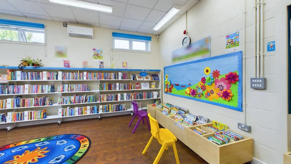 Bright children's library space with shelves of books, colourful chairs, reading display, and educational floor mat.