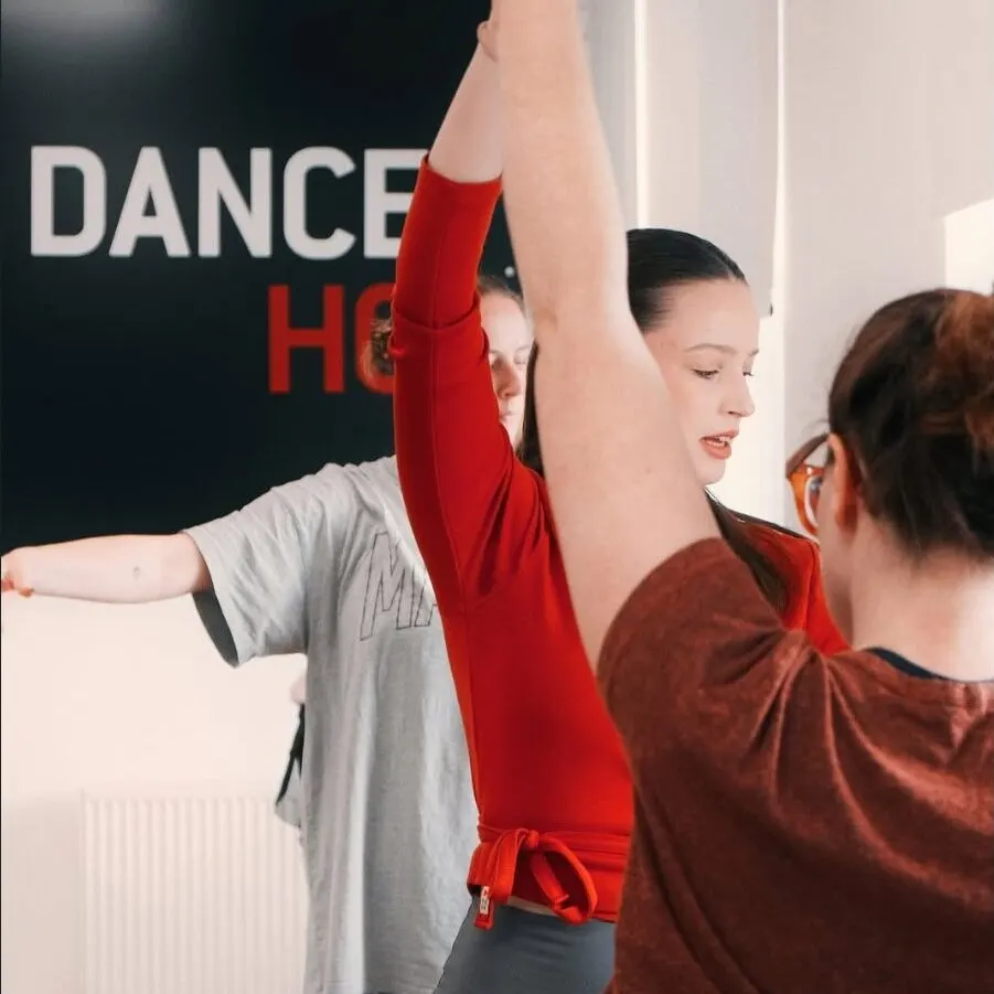 A dancer in red arm sleeves and grey top practises with an instructor during a class at a dance studio.